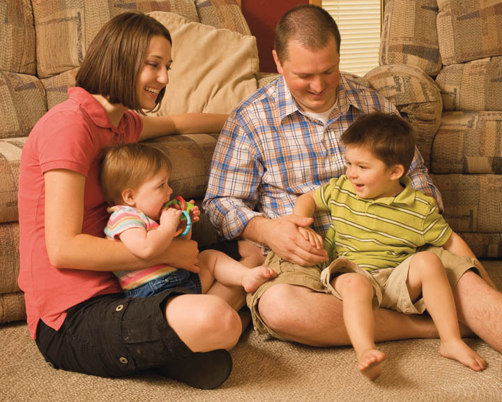 A photo of a Mormon family in their home; consists of a father, a mother, and young children.