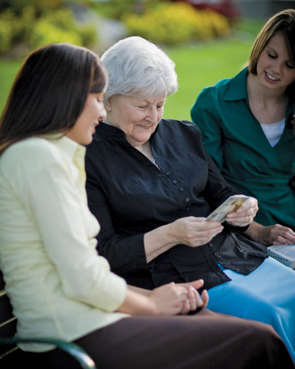 A photo of two Mormon sister missionaries speaking with a woman on a bench. 