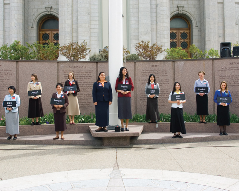 A photo of ten sister missionaries with name badges and signs indicating which language they can speak, standing in front of the Salt Lake Temple, ready to answer questions of passerby's.