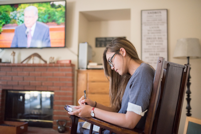 A young woman takes notes while watching general conference at home.